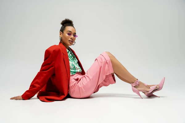 スタイリッシュな服装とグレーの背景に座っているピンクのサングラスの自信を持つアフリカ系アメリカ人女性 — ストック写真
