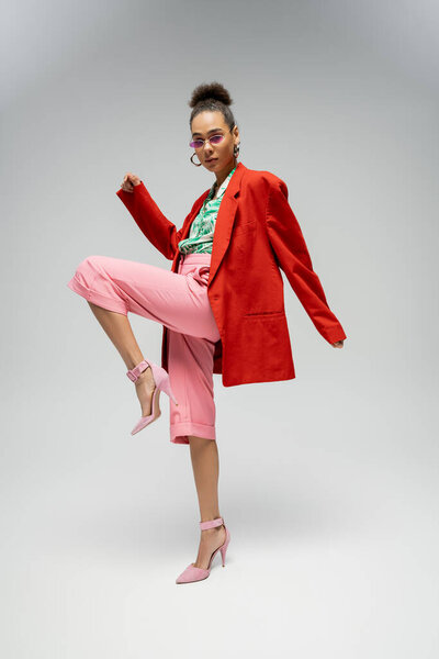 full length of african american model in bold attire and sunglasses on grey backdrop, dynamic pose