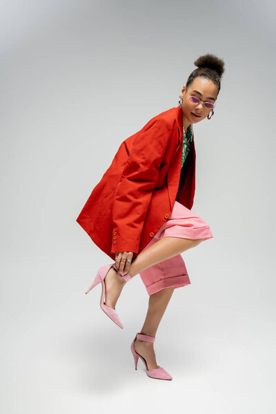 african american fashion model in red blazer adjusting strap on high heels on grey backdrop
