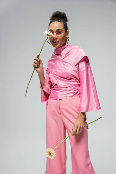 cheerful african american woman in pink attire and sunglasses posing with flowers on grey backdrop