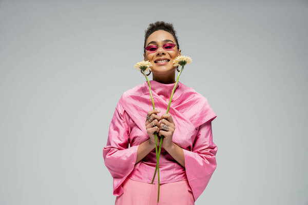 pleased african american woman in pink attire and sunglasses posing with flowers on grey backdrop