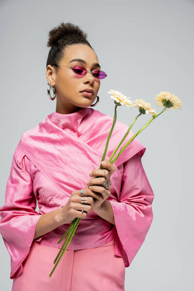 stylish african american woman in pink attire and sunglasses posing with flowers on grey backdrop