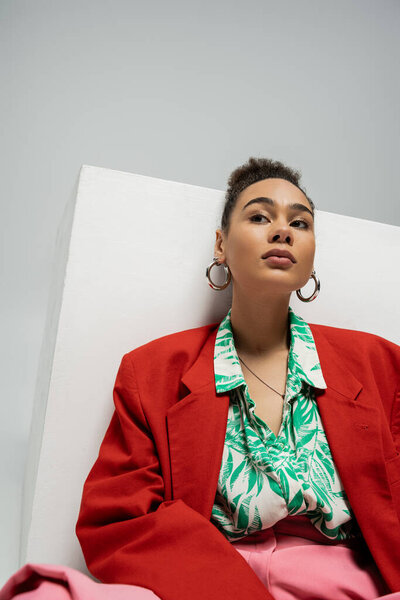 Мечтательная африканская американка в модном и ярком пиджаке позирует рядом с кубом на сером фоне