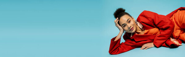 happy african american model in silver accessories and vibrant outfit lying on blue backdrop, banner