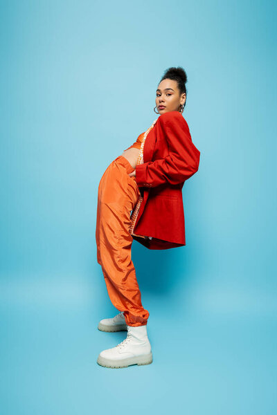 stylish fashion model in orange suit and red blazer posing on blue backdrop and looking at camera