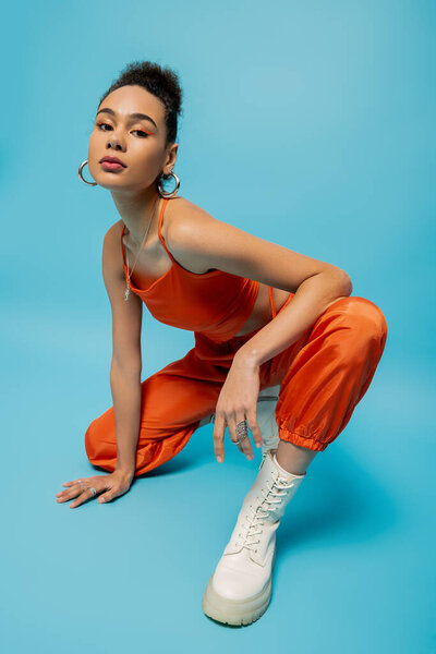 attractive african american model in orange outfit squatted with her hand on floor on blue backdrop