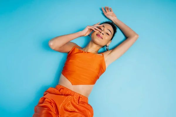 cheerful fashion model in vivid orange outfit lying on blue floor slightly smiling and touching face