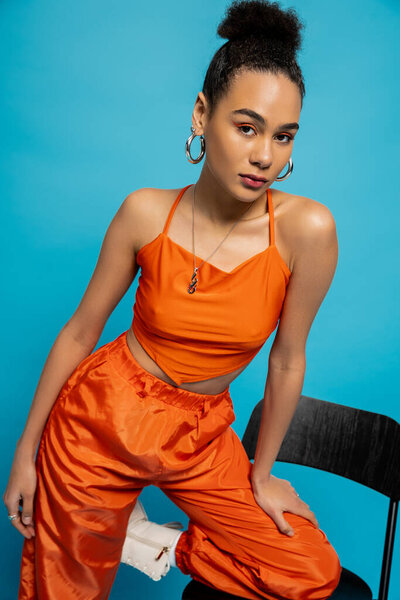 beautiful fashion model in orange outfit posing on tall chair looking at camera, blue background