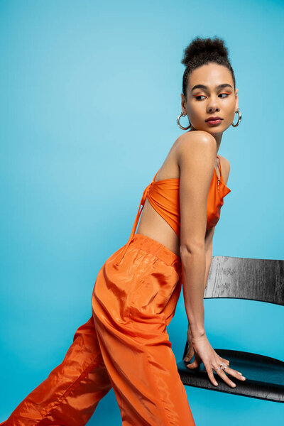 pretty young fashion model in orange stylish attire posing with tall chair on blue backdrop