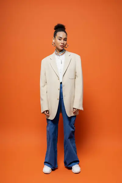 stylish african american woman in urban outfit with beige blazer posing on bright orange backdrop