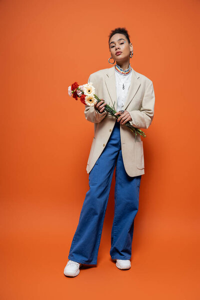 привлекательная модель в повседневной городской одежде, позирующая на оранжевом фоне с цветами в руках