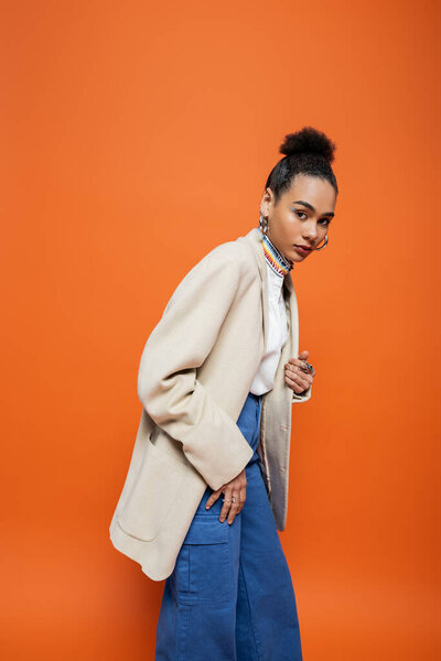 elegant african american woman in beige blazer and blue pants looking at camera, orange backdrop