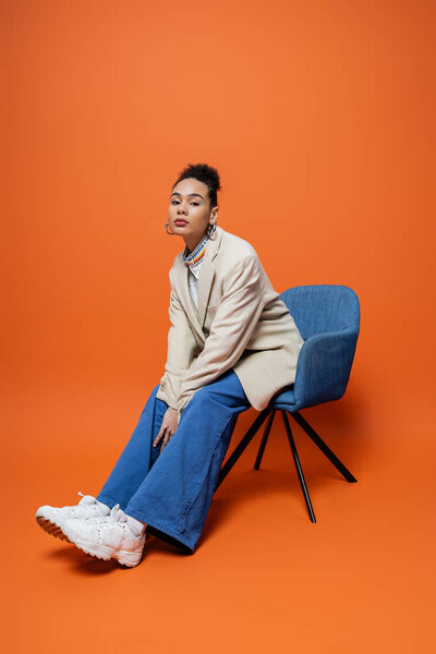 красивая модель в бежевом блейзере синие штаны и яркий макияж сидя на синем кресле