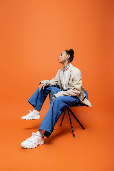 stylish african american model in fashionable and trendy outfit sitting on blue chair looking away