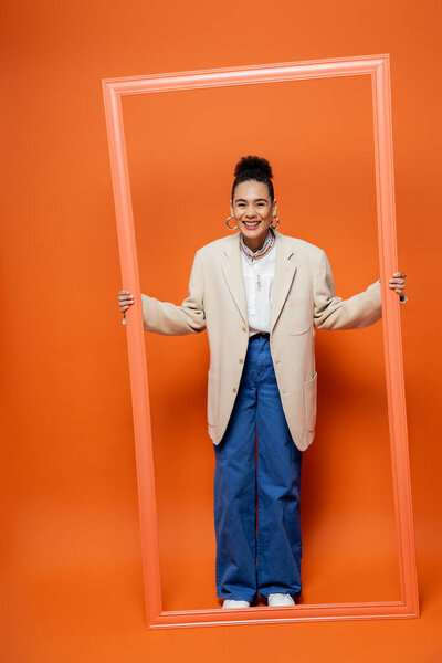 cheerful attractive woman in blazer and blue pants holding orange framework on orange backdrop