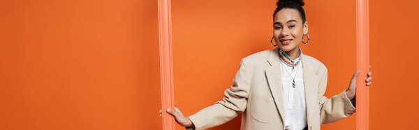 cheerful african american woman in beige blazer with hoop earrings holding orange framework, banner