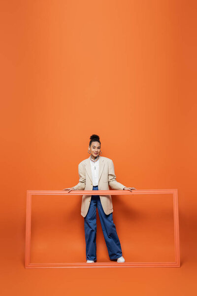 smiling african american model in beige blazer and blue pants holding frame on orange backdrop