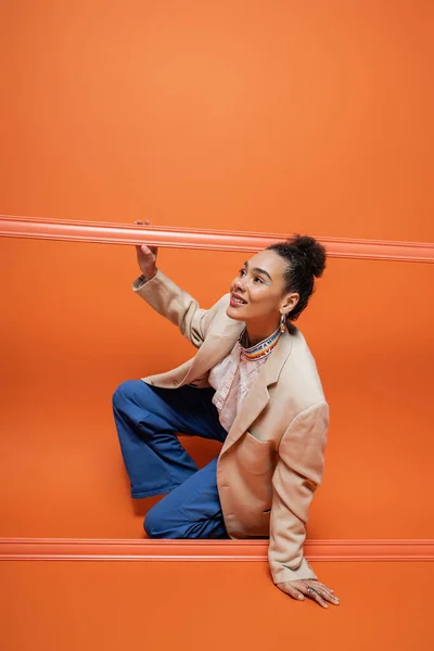 cheerful african american fashion model in stylish outfit squatting out of orange framework