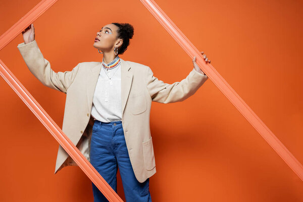 trendy african american model in stylish street attire with accessories looking away holding frame