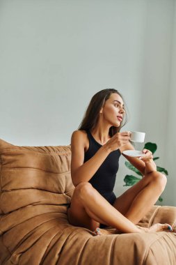 Kumral saçlı güzel genç bir kadın kahve keyfi yapıyor, rahat bir fasulye koltuğu üzerinde oturuyor.