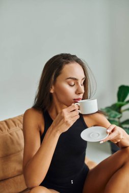 Kumral saçlı güzel genç bir kadın sabah kahvesinin tadını çıkarıyor, fasulye sırığında oturuyor.