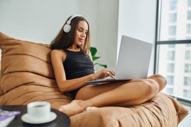 Kablosuz kulaklıklı, dizüstü bilgisayar kullanan ve fasulye koltukta oturan serbest çalışan, güzel bir kadın.