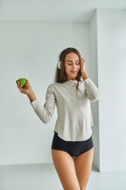 pleased brunette woman in long sleeve and panties listening music in headphones and holding apple clipart