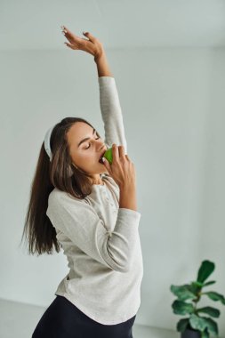 brunette woman in long sleeve listening music in headphones and biting green apple, enjoying taste clipart