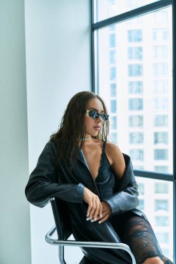 portrait of seductive woman in sunglasses posing in lace underwear and leather coat near window clipart