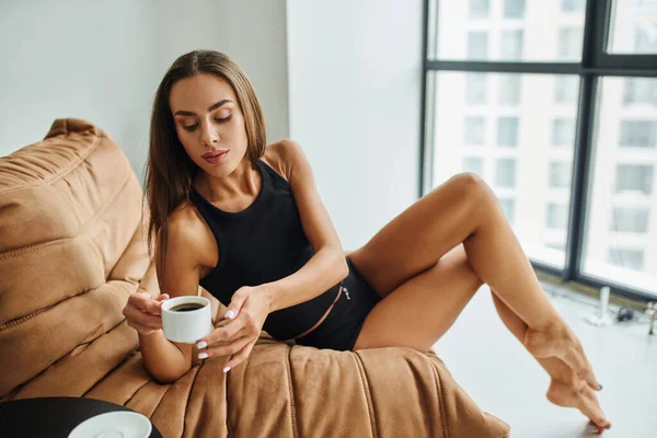 stock image barefoot young woman with brunette hair sitting on bean bag chair and reaching cup of black coffee