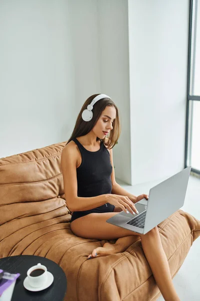 barefoot freelancer in wireless headphones using laptop and sitting on bean bag chair, pretty woman