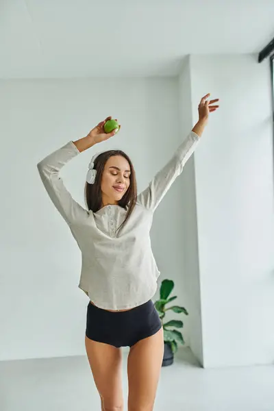 happy brunette woman in long sleeve and panties dancing while listening music in headphones
