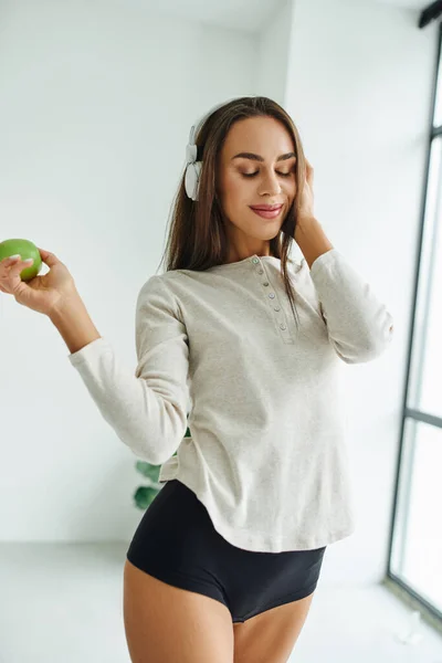 stock image joyful woman in long sleeve and panties listening music in headphones and holding apple
