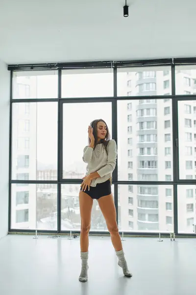 young woman in long sleeve and panties listening music in headphones near panoramic windows