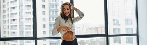 счастливая женщина показывает пирсинг живота и прослушивания музыки в наушниках возле окон дома, баннер