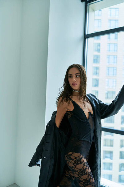 passionate woman with brunette hair posing in leather coat and black lace underwear near window