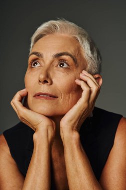 portrait of dreamy senior woman with short silver hair looking away with hands near face on grey clipart