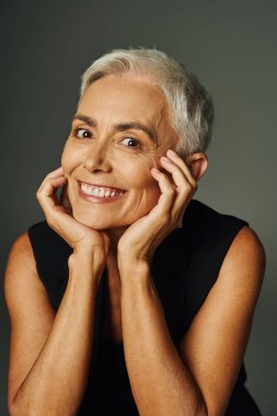 portrait of joyful senior lady with short silver hair looking at camera on grey, elegance and charm clipart