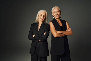 Gülümseyen siyah şık elbiseli yaşlı modeller gri, eski moda kollarını kavuşturmuş duruyorlar.