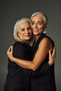 lifelong friendship, happy and trendy senior women in black attire embracing on grey backdrop clipart