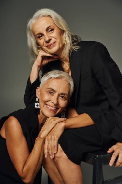 joyful senior models posing in black stylish attire on grey, elegant aging of lifelong friends clipart