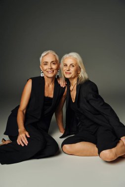smiling senior models in black stylish attire sitting on grey backdrop, elegant aging and friendship clipart