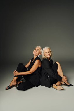 cheerful senior female friends in black attire sitting back to back on grey, fashionable aging clipart