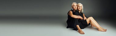 fashionable barefoot senior models in black casual attire sitting on grey backdrop, banner clipart