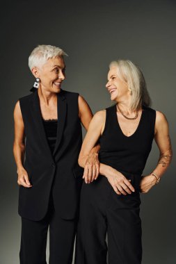 laughing senior women in black trendy attire looking at each other on grey, lifelong friends clipart