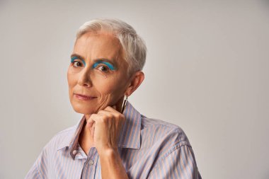 fashionable senior model with bold makeup and short silver hair looking at camera on grey, banner clipart