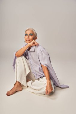 barefoot and dreamy senior model in blue striped shirt and pants sitting and looking away on grey clipart