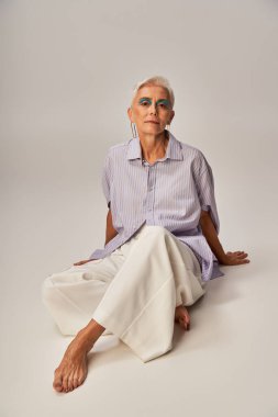 barefoot and confident senior lady in trendy casual attire looking at camera while sitting on grey clipart