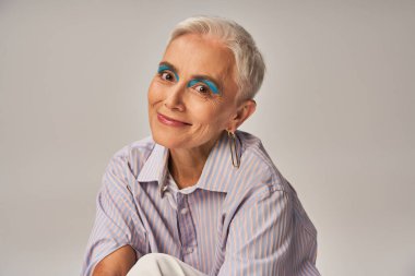 positive mature lady with bold makeup in blue striped shirt smiling at camera on grey, banner clipart