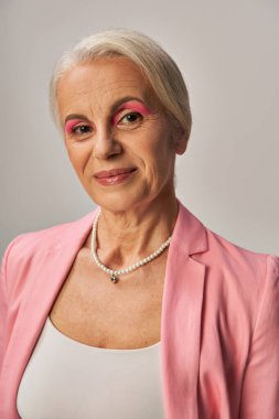 portrait of elegant senior lady in pink blazer and pearl necklace smiling at camera on grey clipart